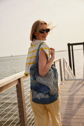 Model posing and wearing light white and yellow clothes and blue net beach bag.