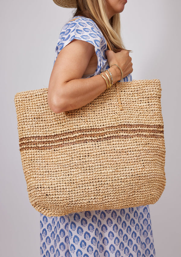 Model wearing blue kaftan and holding straw bag with brown stripes