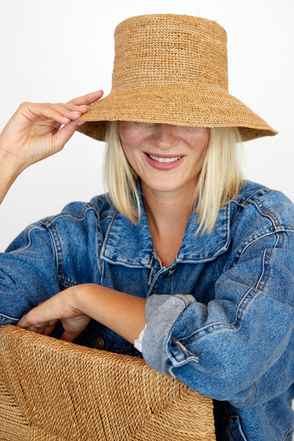 Model holding brim of straw bucket hat and wearing denim shirt