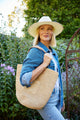 Model holding a straw tote with leather handles and wearing a sunhat