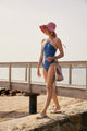 Model wearing blue bathing suit, pink canvas sunhat and holding a colorful tote