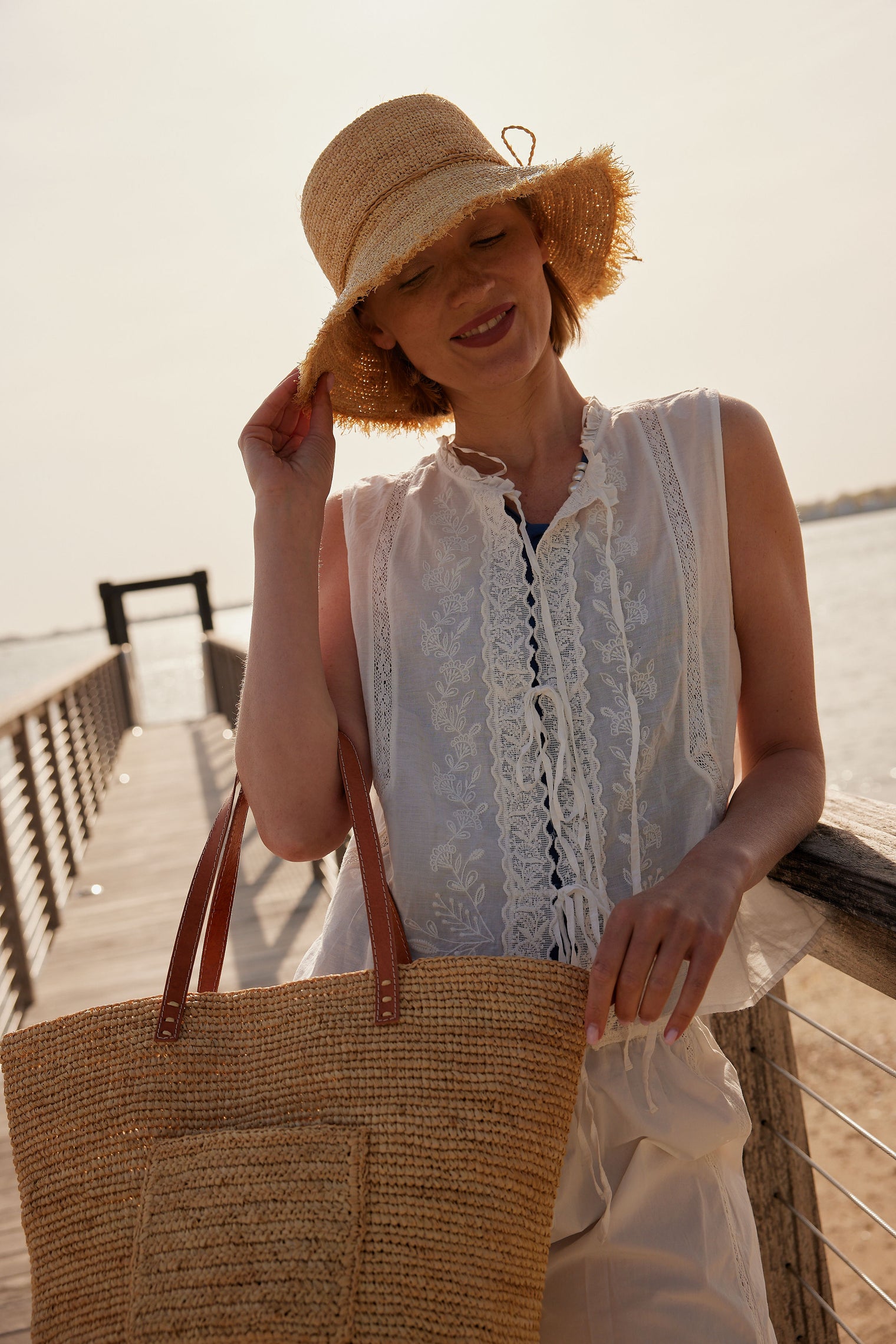 Model wearing a frayed edge straw raffia bucket hat and holding a straw tote on a pier