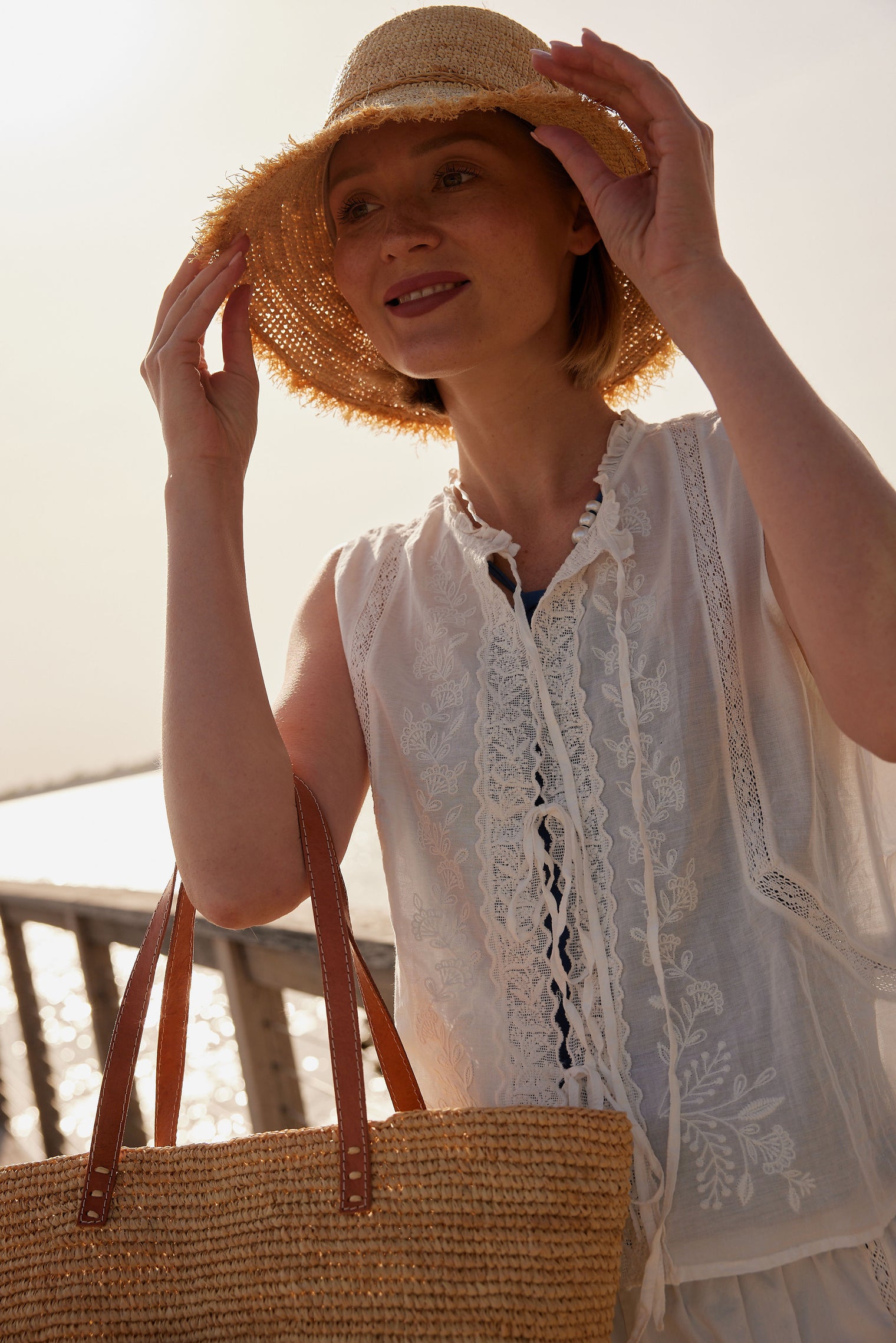 Model holding a brim of a frayed edge straw raffia bucket hat on a pier