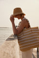 Model wearing white striped straw tote bag and holding brim of straw bucket hat