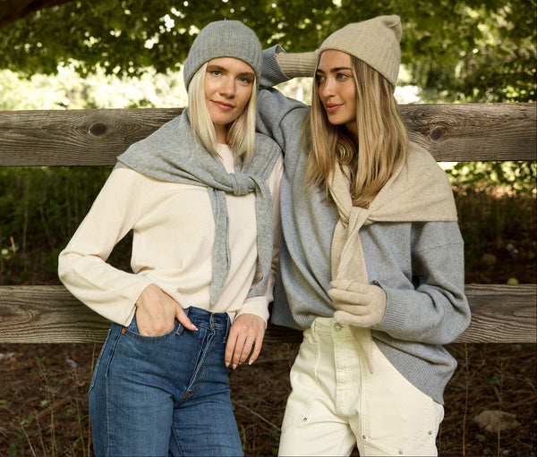 Two women posing outdoors against a rustic wooden fence, wearing coordinated knit sweaters, scarves, and beanies in neutral cream, beige, and gray tones.