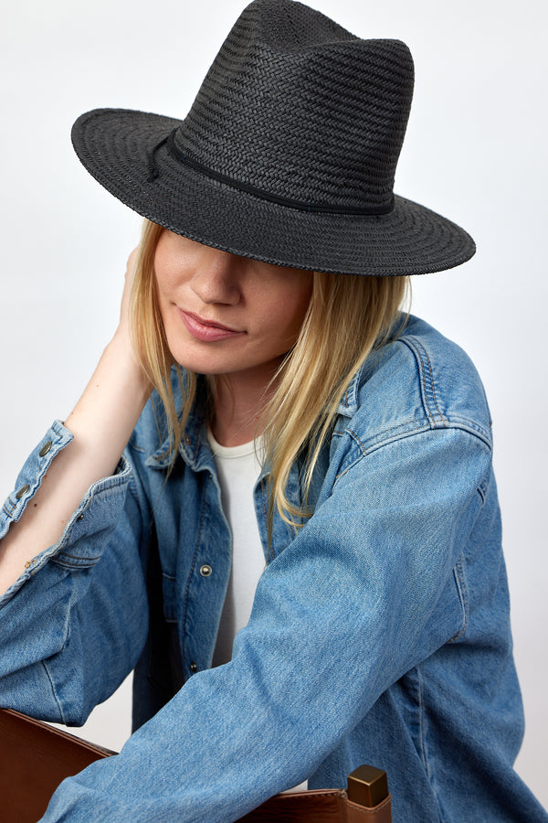 Model wearing black straw fedora and denim shirt while sitting in chair