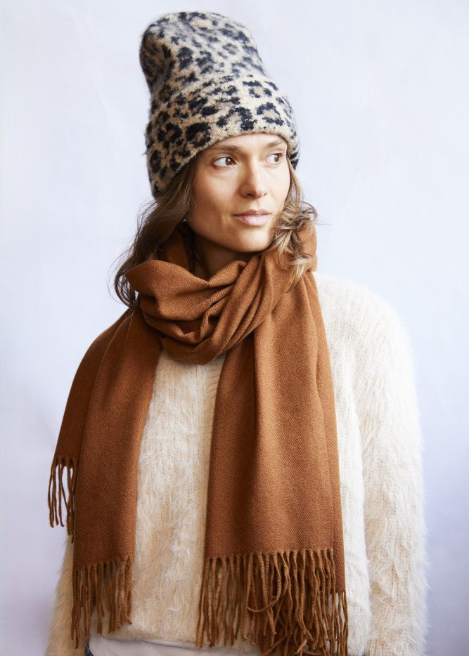 Woman wearing a leopard print beanie and brown scarf against a white background