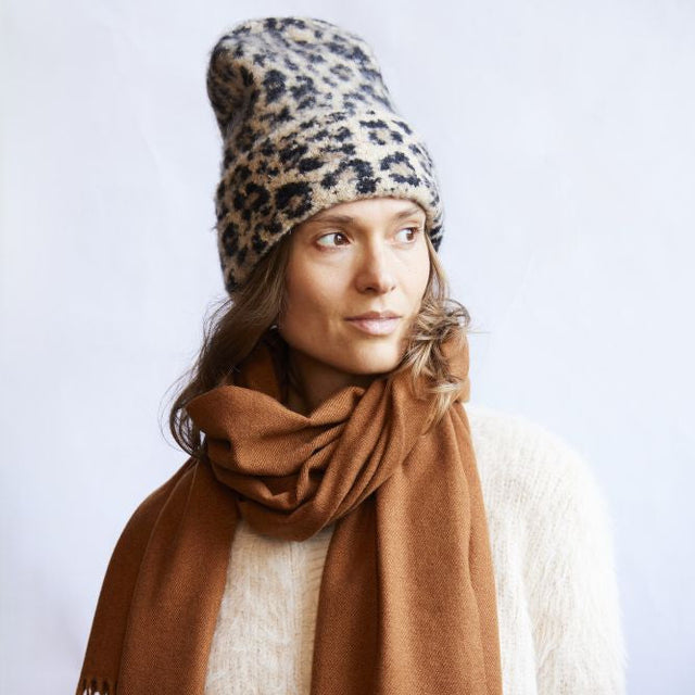 Woman wearing a leopard print beanie and brown scarf against a white background