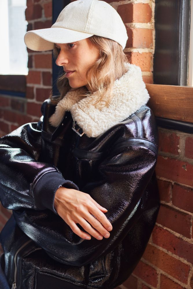 Woman wearing a dark leather jacket with faux fur lining and a cream cap against a brick wall.
