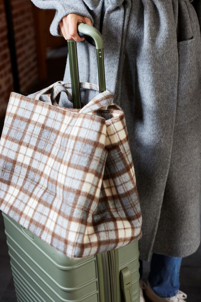Person holding a plaid bag over a green suitcase with a gray coat and blue jeans.