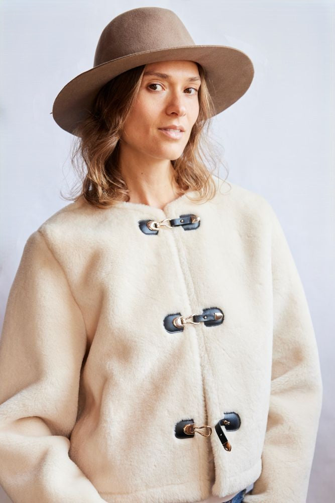 Woman wearing a beige coat with black buttons and a brown hat on a light gray background