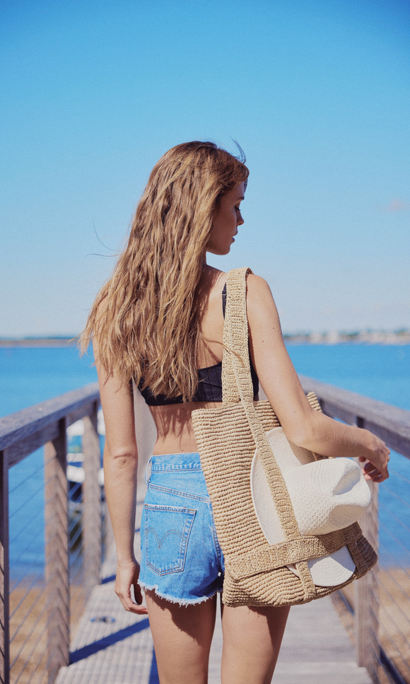 A woman with long, wavy hair, wearing a black top and denim shorts, walks on a wooden pier by the water, carrying a straw bag and a hat.