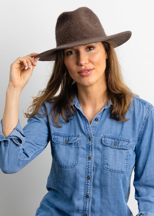 Model holding brim of brown velour brimmed hat and wearing denim shirt