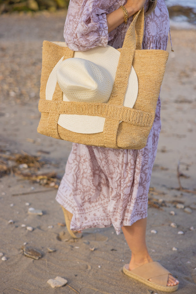 Model holding straw bag with straps that hold a sun hat walking in the sand