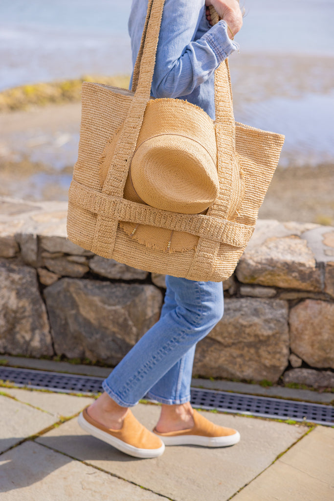 Model holding straw bag with straps that hold a sun hat outside by the beach