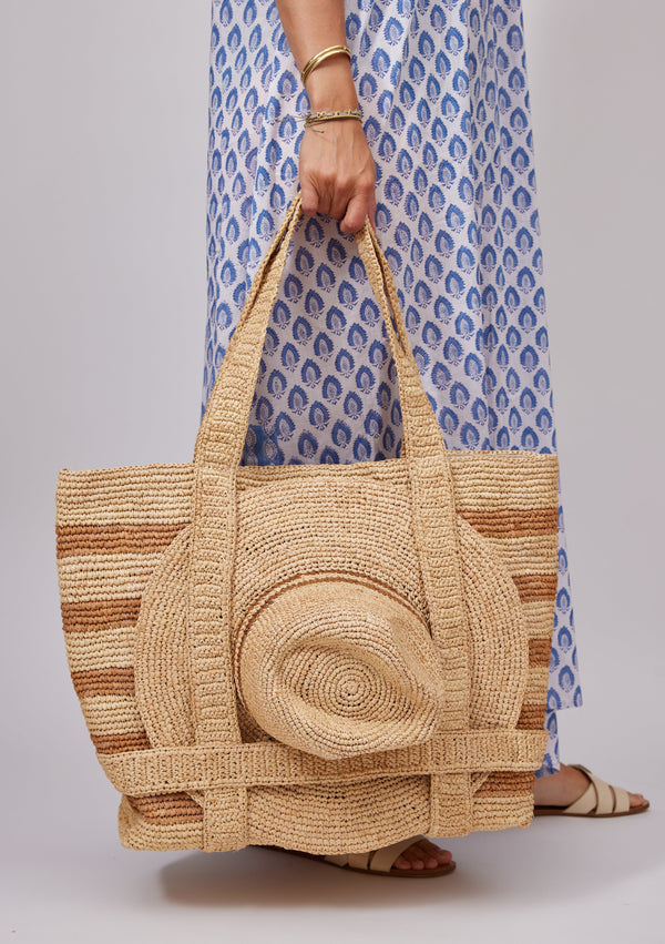 Model wearing a blue kaftan and holding a tan striped straw tote with a sun hat attached under straps