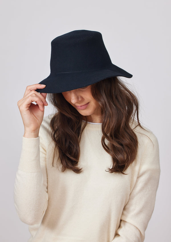 Model holding brim of black velour brimmed hat while wearing it