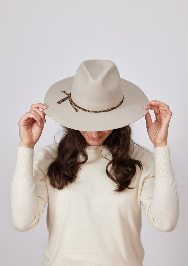 Model holding brim while wearing beige felt brimmed hat with brown chain trim detail