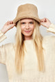 Model wearing ivory sweater and holding brim of tan faux fur bucket hat