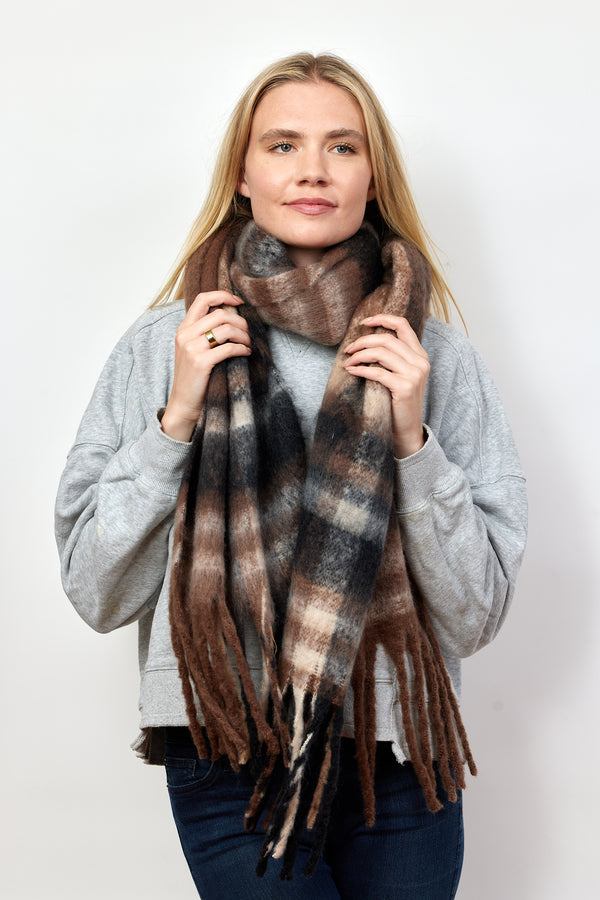 Model holding black, brown and tan plaid scarf with fringe detail around neck