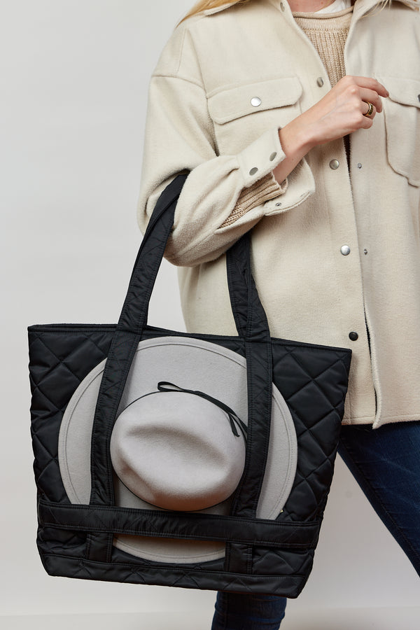 Model holding black quilted nylon bag with straps to hold a hat and wearing tan jacket