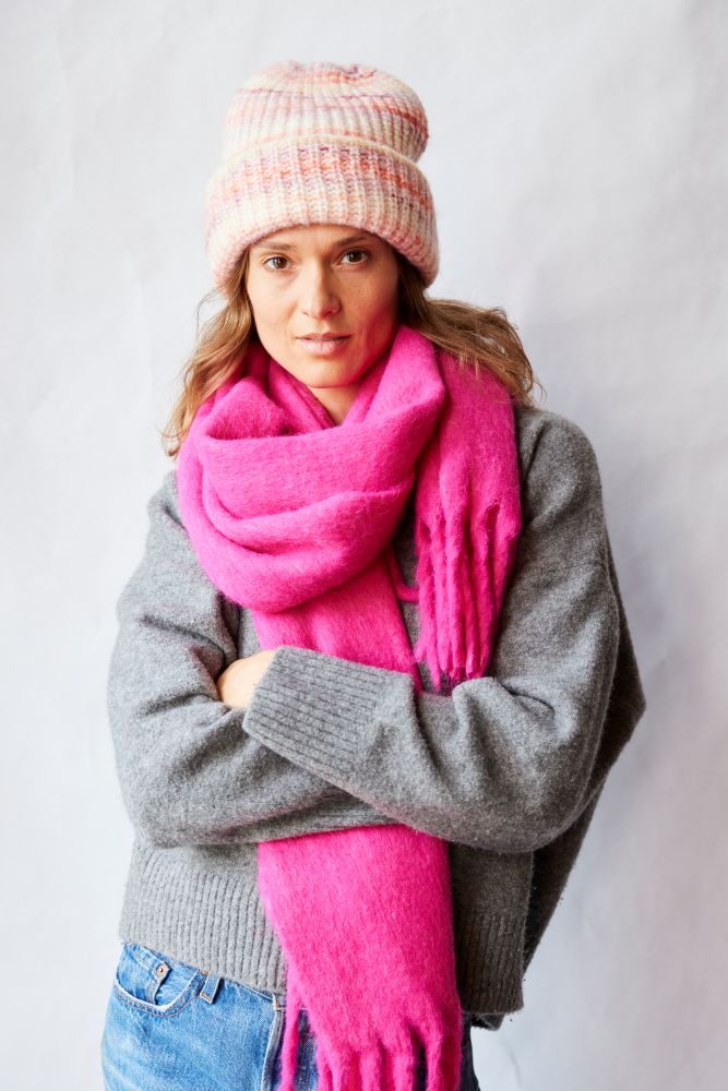 Woman wearing a pink scarf, gray sweater, and striped beanie against a white background