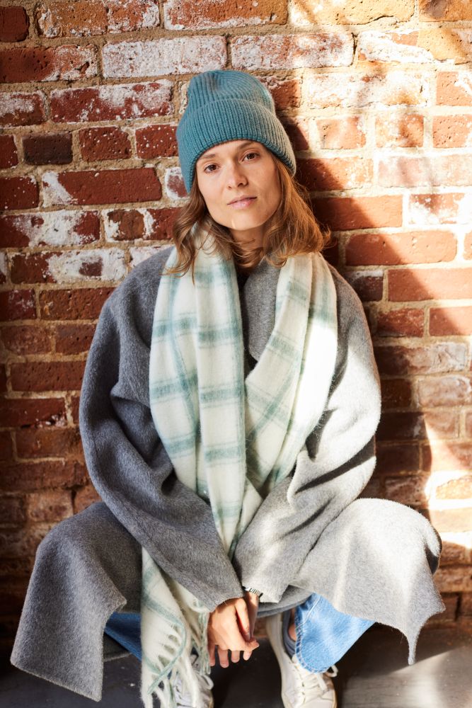 Person wearing a blue beanie, gray coat, and plaid scarf against a brick wall.