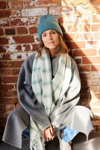 Person wearing a blue beanie, gray coat, and plaid scarf against a brick wall.