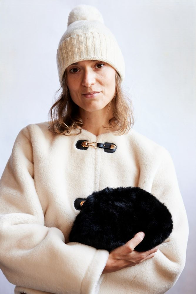 Woman wearing a cream coat and white beanie holding a black fur clutch against a white background