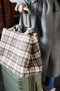Person holding a plaid bag over a green suitcase with a gray coat and blue jeans.