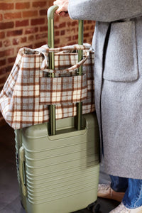 Person holding a green suitcase with a plaid bag on top, against a brick wall.