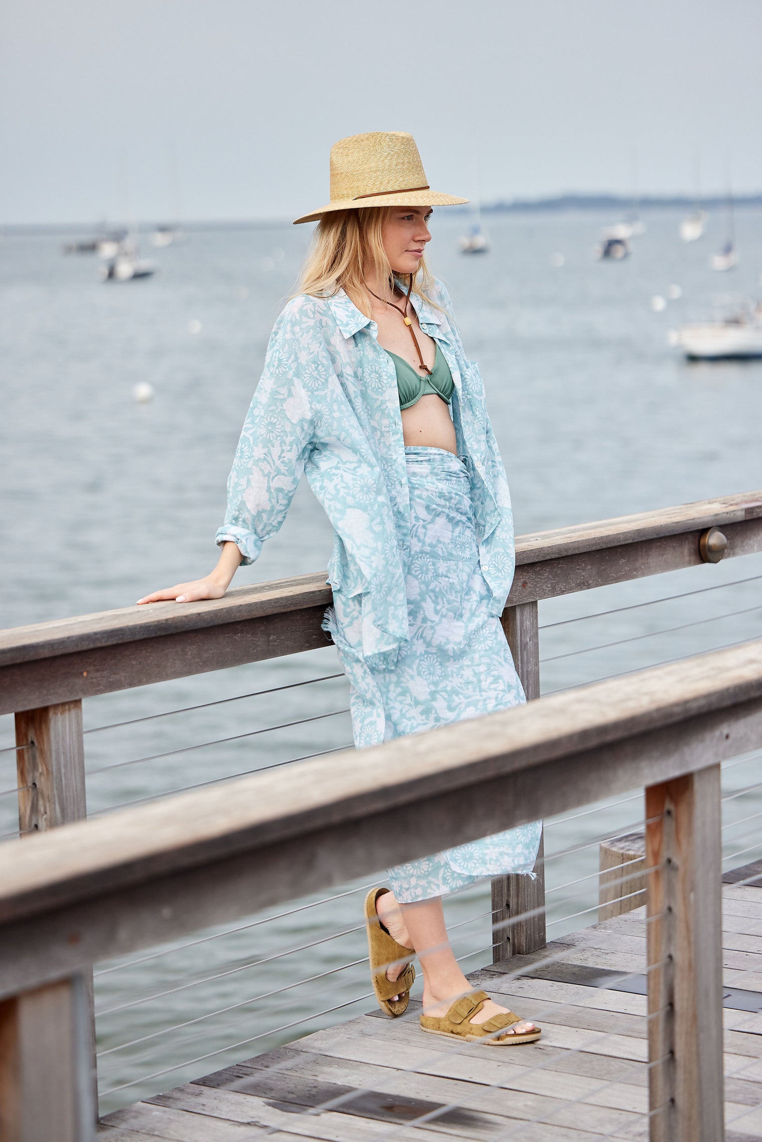 Woman in a light blue outfit and straw hat standing on a wooden pier by the water.