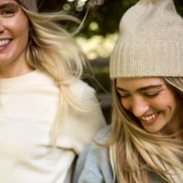 Two women smiling outdoors in cozy fall/winter attire, including knit beanies, sweaters, and a pair of taupe knit gloves. One woman wears a cream sweater and jeans, while the other wears a light blue sweater, a beige beanie, and taupe gloves.